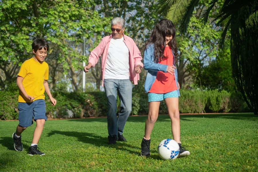 Een oudere man en twee kinderen spelen met een voetbal op gras